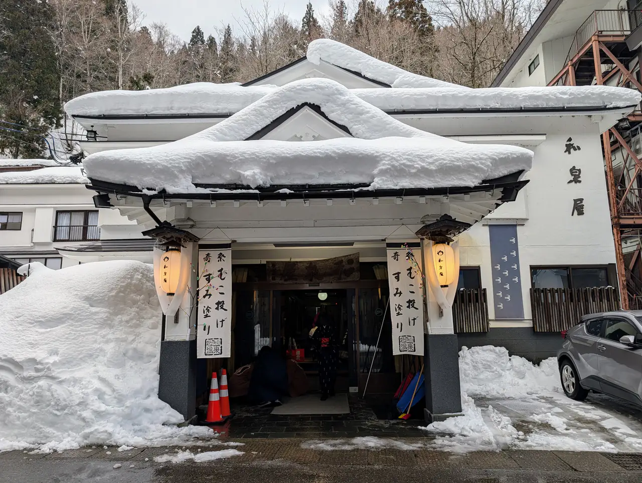 A ryokan entrance in winter: a snow-capped gabled porch with paper lanterns and hanging signs, deep snow banked on either side