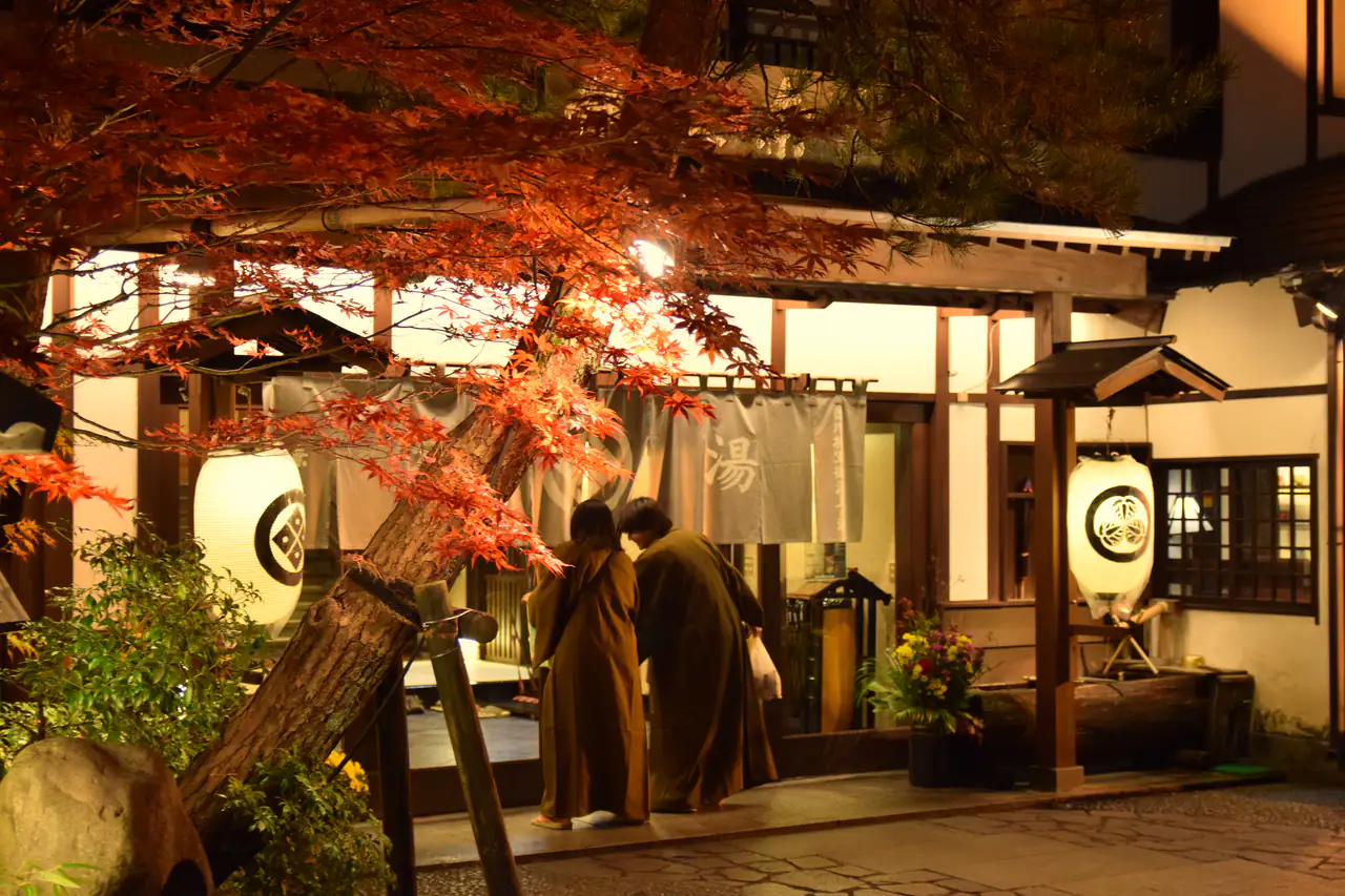 At night, people walk toward a lantern-lit wooden bathhouse entrance framed by bright red maple leaves