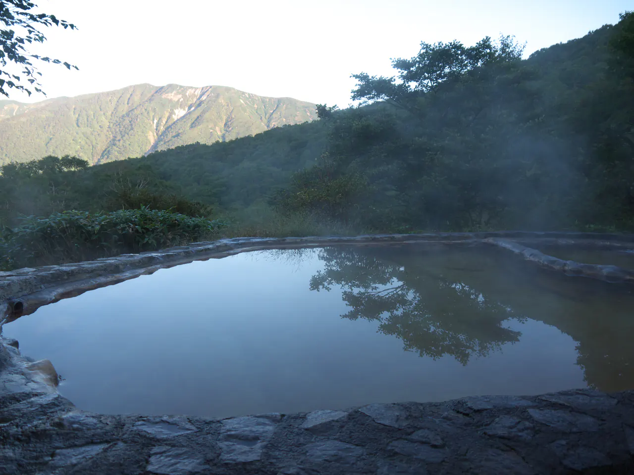 A simple stone-edged outdoor pool reflecting the sky, with forested mountain ridges and drifting steam beyond