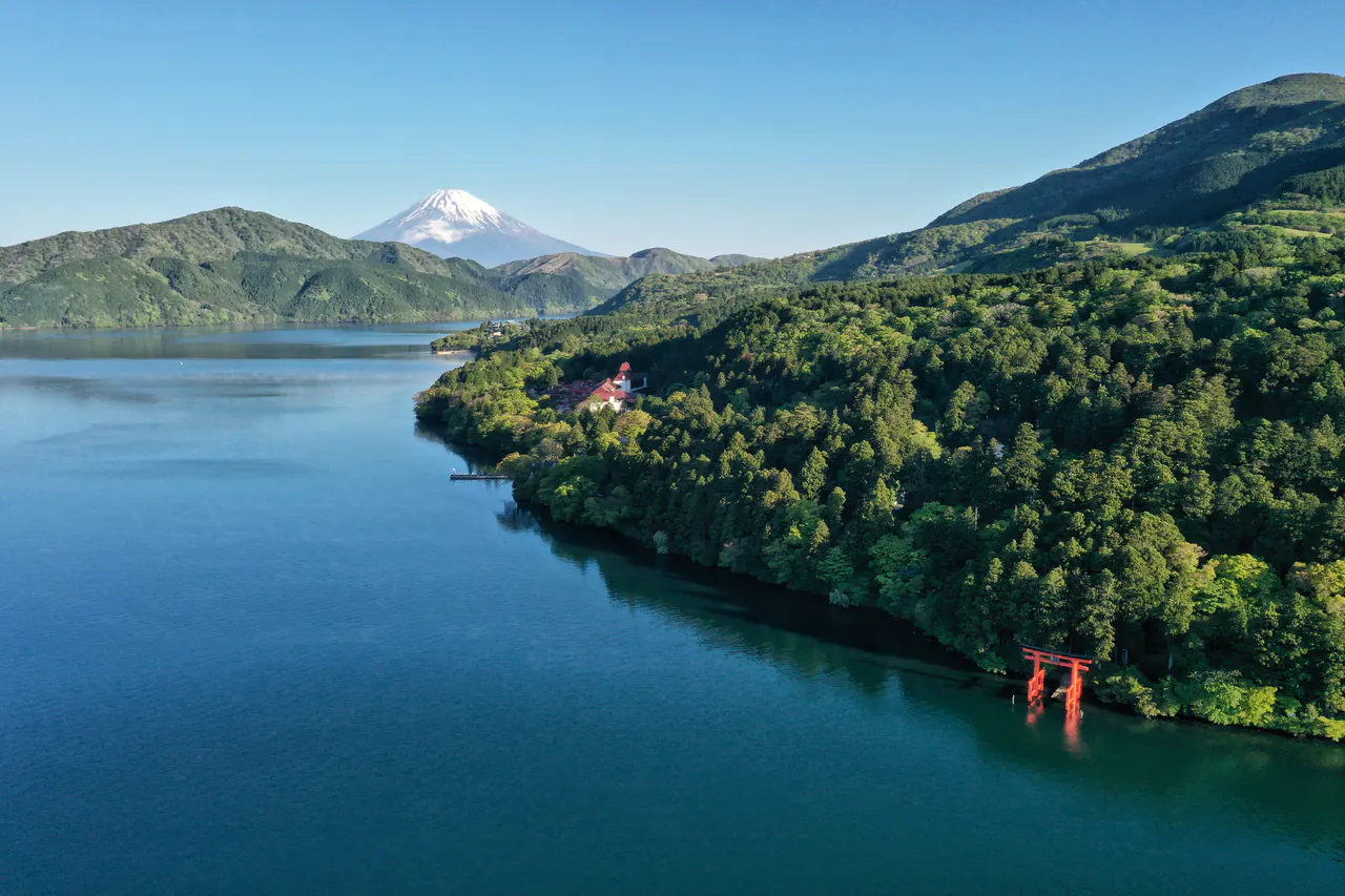 Mount Fuji capped with snow above the deep blue Lake Ashi, forested hills and a red torii gate standing in the water
