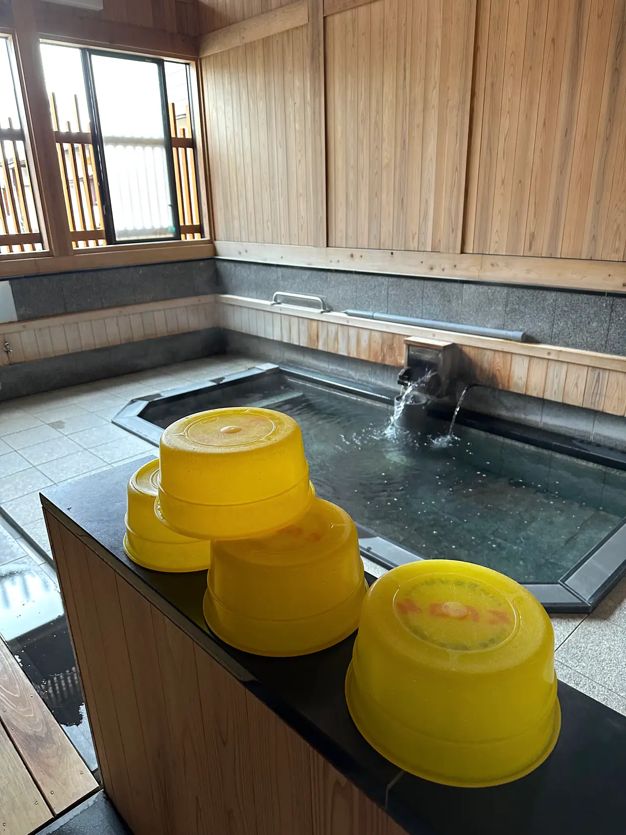 A simple wooden communal bath with a steaming tub, yellow plastic washbowls stacked on the ledge in the foreground