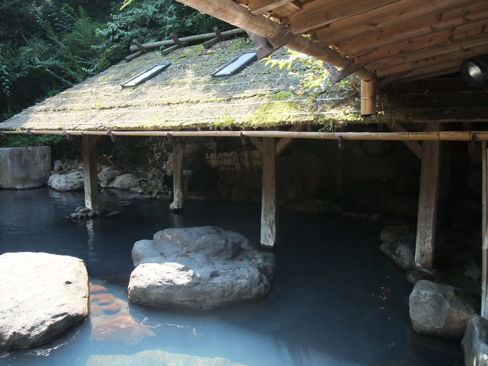 A quiet outdoor bath of pale blue water among large stones, sheltered by a moss-covered wooden roof in the forest