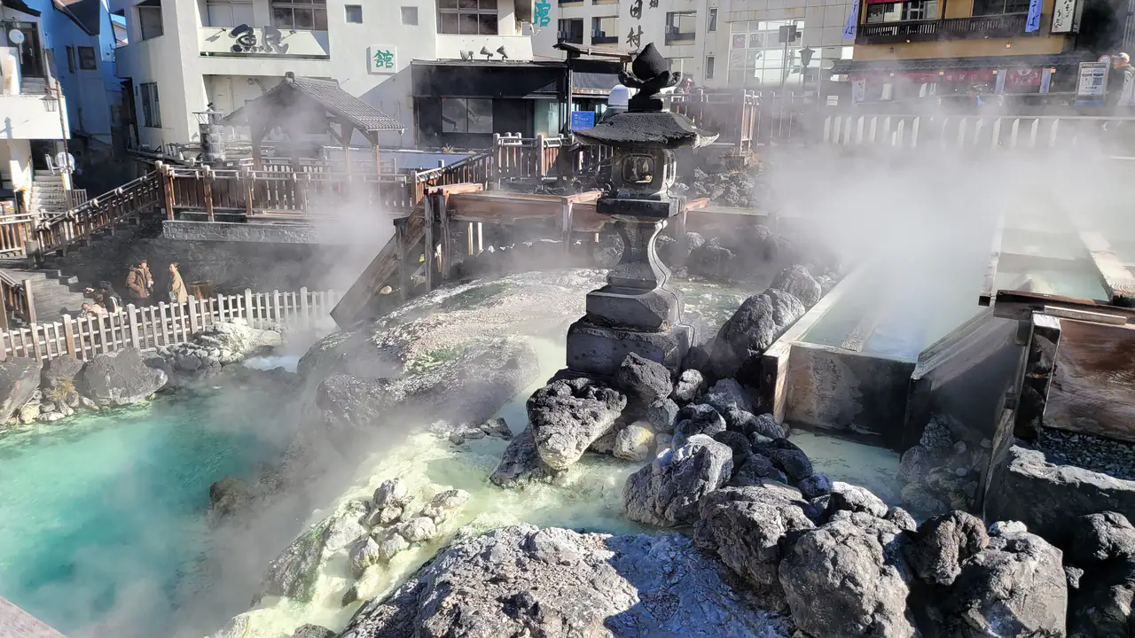 Clouds of steam rise over turquoise spring water and dark volcanic rocks at the Yubatake in Kusatsu, with a stone lantern beside wooden water channels