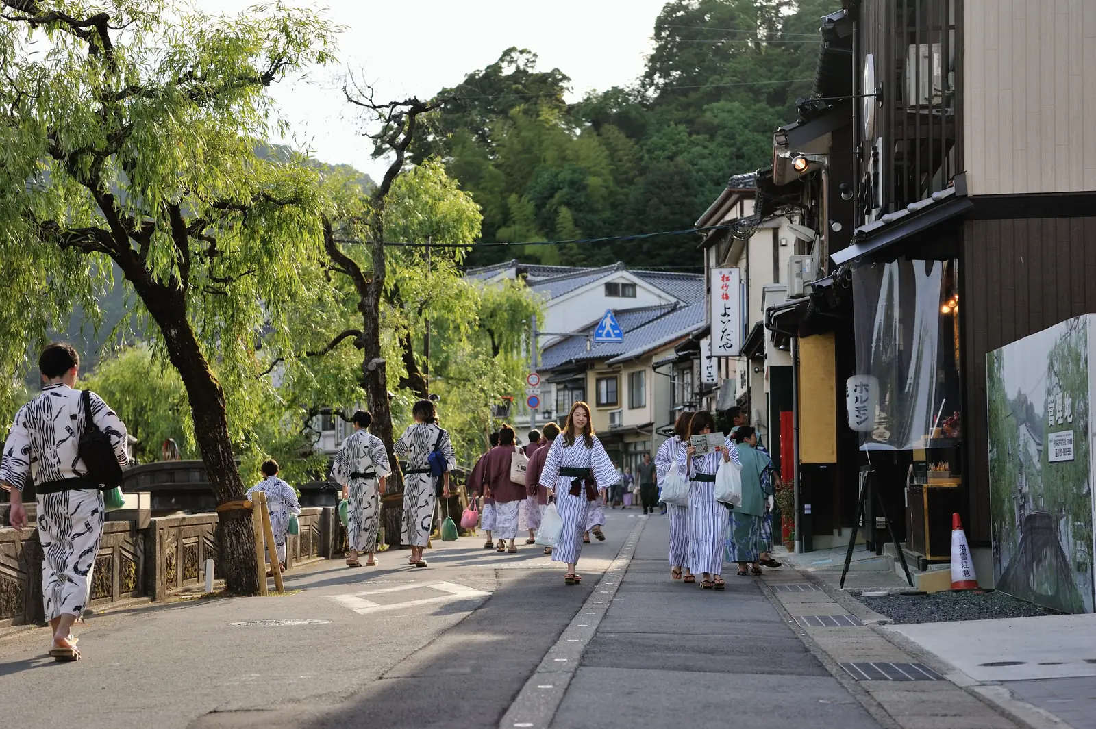 Guests in striped ryokan yukata stroll along a willow-lined street in Kinosaki Onsen on a summer evening