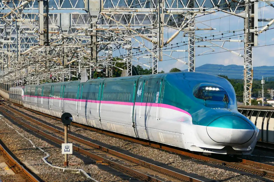 A green and white E5 series Hayabusa Shinkansen with a pink stripe and a long nose arrives under platform roofs